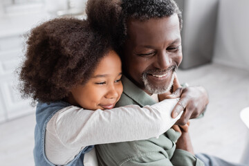 happy african american girl with curly hair hugging cheerful grandfather at home.