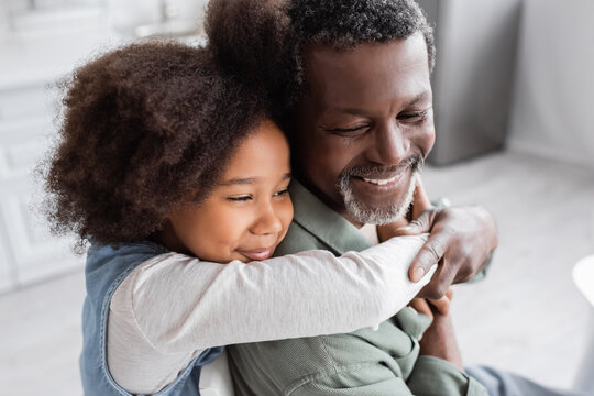 happy african american girl with curly hair hugging cheerful grandfather at home.