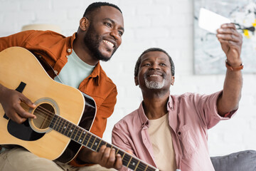 Smiling african american father taking selfie with son playing acoustic guitar at home.