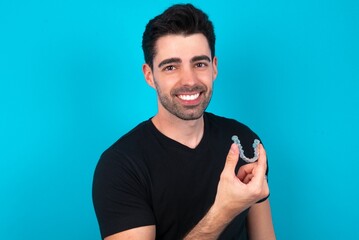 Young man wearing black T-shirt over blue studio background holding an invisible aligner ready to use it. Dental healthcare and confidence concept.