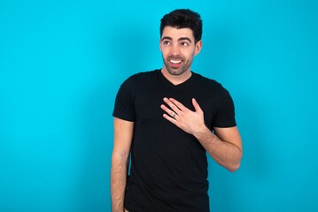 Fototapeta premium Joyful Young man wearing black T-shirt over blue studio background expresses positive emotions recalls something funny keeps hand on chest and giggles happily.