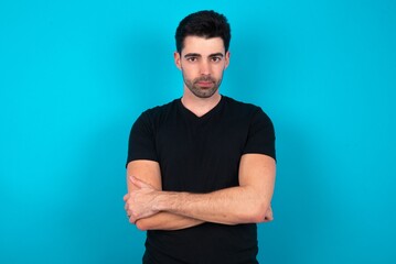 Self confident serious calm Young man wearing black T-shirt over blue studio background stands with arms folded. Shows professional vibe stands in assertive pose.