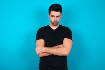 Fototapeta premium Gloomy dissatisfied Young man wearing black T-shirt over blue studio background looks with miserable expression at camera from under forehead, makes unhappy grimace