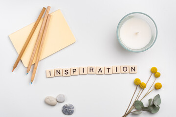 Clean workspace with pencils and notebook, candle and flowers on white background. The word INSPIRATION written with tile letters. Flat lay