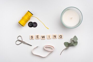 flat lay of scissors, yellow thread roll, buttons and candle on white background. word SEWING made with tile letters 