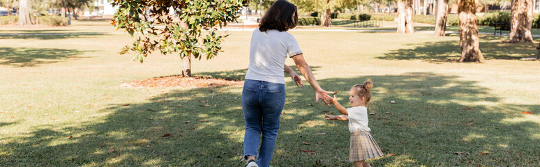 brunette mother in jeans playing with happy toddler daughter in park of Miami, banner.