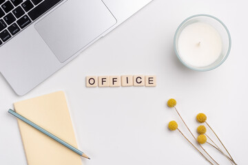 Clean workspace with laptop, pencil and notebook, candle and flowers on white background. The word OFFICE written with tile letters. Flat lay