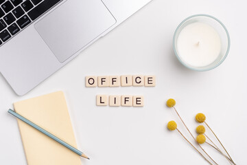 Clean workspace with laptop, pencil and notebook, candle and flowers on white background. OFFICE LIFE written with tile letters. Flat lay