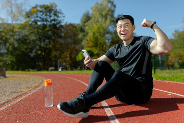 Portrait of a young Asian man, an athlete sitting on a treadmill with a phone. He is happy, looks into the camera. Shows a gesture of victory and strength. Checks the results of running, training.