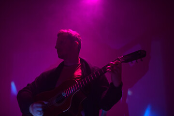 Musician playing acoustic guitar in a foggy club with colorful lights.