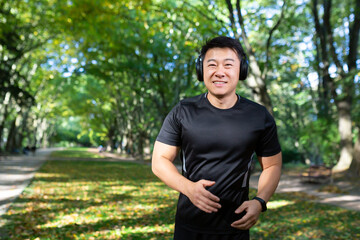 Portrait of a young Asian male athlete running in the park, doing a morning jog in headphones, listening to music. audiobook, podcast. He looks at the camera, smiles.