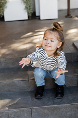 baby girl in casual clothes demanding attention and gesturing while sitting on stairs near house.