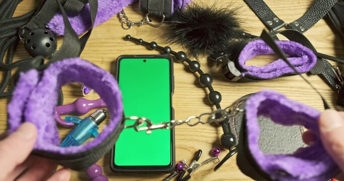Sex toys for erotic games, soft handcuffs, smartphone with green screen. Men's hands put handcuffs on table.