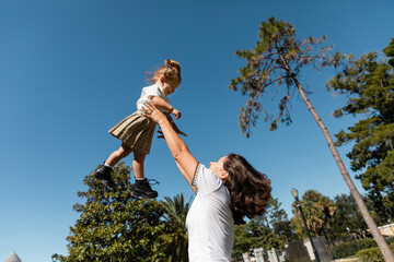 happy mother lifting toddler daughter against green trees and clear sky.