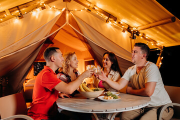 Happy group of friends relaxing in glamping and drinking wine on summer evening near cozy bonfire. Luxury camping tent for outdoor recreation and recreation. Lifestyle concept - 583587160