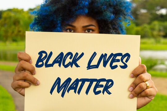 Uma mulher jovem, com cabelos pintados de azul, com rosto escondido atr&aacute;s de um cartaz com o texto: "Vidas Negras Importam".