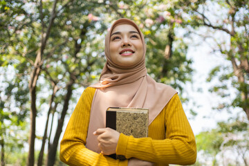 Fototapeta premium Portrait of a delighted young Asian Muslim girl sitting on a grass at the park, reading a book.