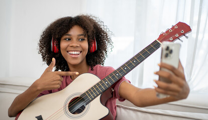 Focused African girl playing acoustic guitar and taking selfie using smartphone while sitting on...