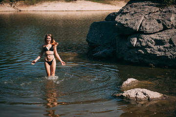 A young girl with long blond hair, a slender figure, in a black swimsuit jumps in the river against the backdrop of rocks and laughs.