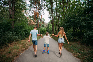 Obraz premium A happy young family, dad, mom and son are skating hand in hand with their parents, on roller skates, along a path in the park. The whole family is dressed in shorts and T-shirts.