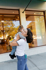 happy mother hugging baby girl in striped long sleeve shirt near showcase of shop in Miami.