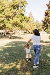 brunette mother holding hands with happy toddler daughter while playing in park of Miami.