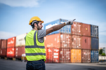 Caucasian warehouse worker in uniform with hard hat using mobile phone standing in container port...