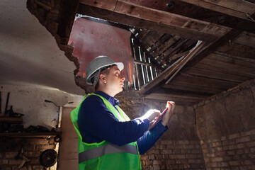 Building inspector. Man in a hard hat and a yellow reflective vest examines damaged structures and...