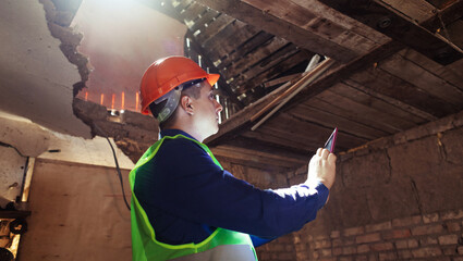 Building inspector. Man in a hard hat and a yellow reflective vest examines damaged structures and...