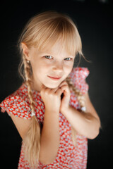 Obraz na płótnie Canvas Portrait of an 8-year-old blonde girl with two pigtails, in a summer floral dress on a dark background, smiling and looking at the camera.