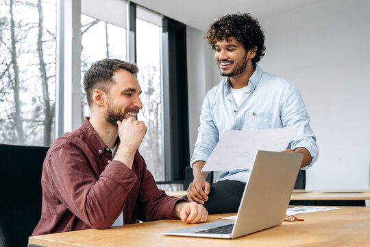 Two positive stylish male business partners of different ethnicity, working together in the office on a new project, brainstorming, discuss ideas and strategy, smile.Collaboration, partnership concept