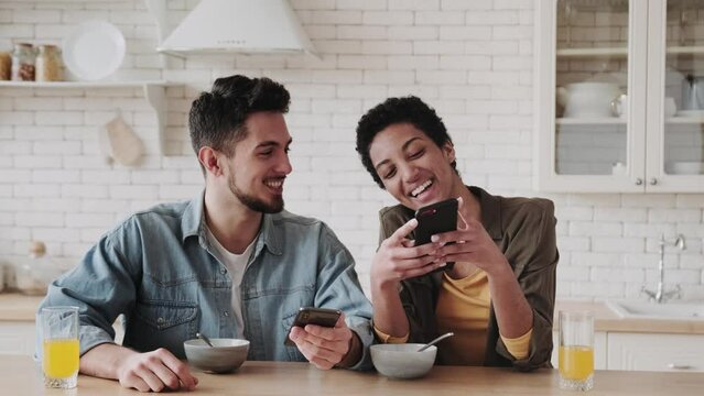Happy young multiethnic couple looking at smartphone and smiling at breakfast in kitchen