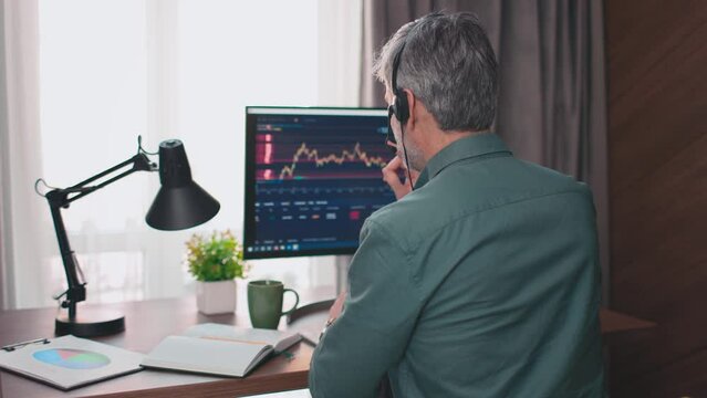 Rear of businessman talking in headset about stock market. Man broker communicating with clients about selling and buing, digital currency and stocks. Working with graphics and charts. Back view.