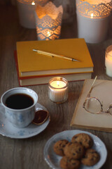 Cup of tea or coffee, plate of cookies, books, e-reader, pencil and lit candles on the table. Selective focus.