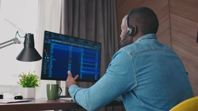 Rear of businessman talking in headset about stock market. African American man broker communicating with clients about selling and buing, digital currency and stocks. Working with graphics and charts