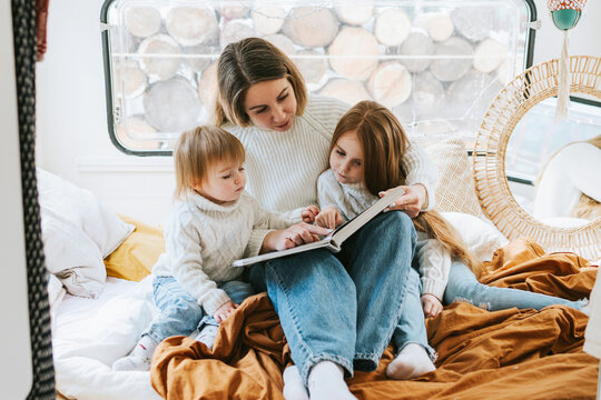 family mom and two red haired cute girl little daughters having fun and reading book in cosy bed in trailer mobile home or recreational vehicle during family local travel