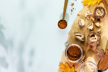 Superfood. Pieces of dry mushrooms with chaga powder in a glass jar on a wooden podium with autumn leaves on a blue background with shadows and copy space. Home herbal apothecary concept. Flat lay