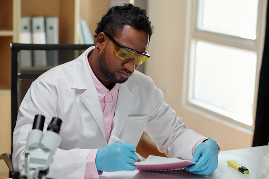 Young man in lab coat and gloves making notes in copybook while sitting by workplace and carrying out new scientific investigation