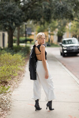 full length of blonde woman in white cargo pants and boots standing with black leather jacket on street in Miami.