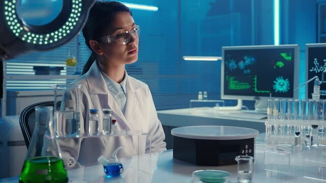 A female Korean doctor examines virtual graphics, zoom and swipe the picture. Asian woman researcher in white gown sits in laboratory with blue light. Biochemical research laboratory. Close up.