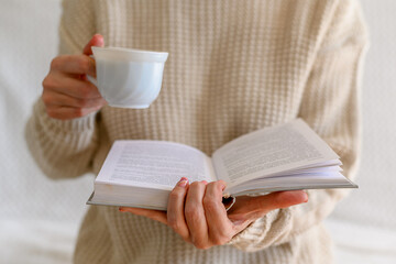 A woman in a white jumper is holding a book and a cup of white coffee.