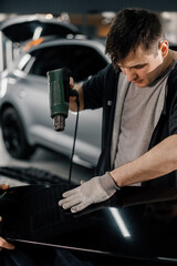 A worker covers a car spoiler with a film in the workshop using a hair dryer and a spatula close-up...