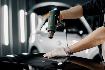A worker covers a car spoiler with a film in the workshop using a hair dryer and a spatula close-up Car detailing and care