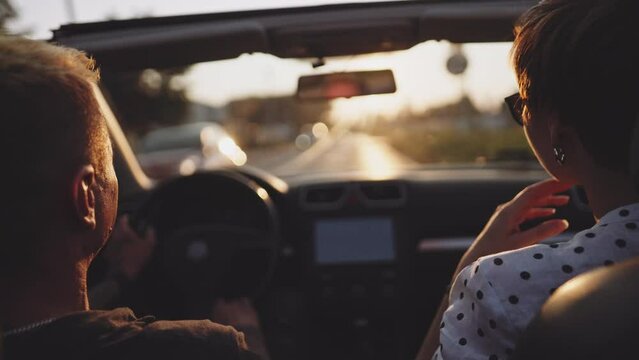 Couple driving and talking in a convertible car at summer sunset. Romantic man and woman in love enjoying warm evening, having a ride in a countryside in a convertible, lens flare sunny view. 