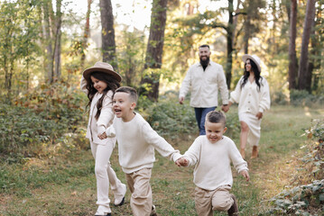 Fototapeta premium Young mother, father with daughter and sons are walking, having fun in autumn forest. Family holding hands enjoying time together at background of trees. Happy parenthood and childhood concept.