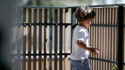 Fototapeta premium Child standing by balcony fence looking outdoors from second floor. Child holding in wooden bars staring outside