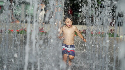 Little boy running at water jets in city park during summer day. Small child runs in refreshing water splashing in slow motion