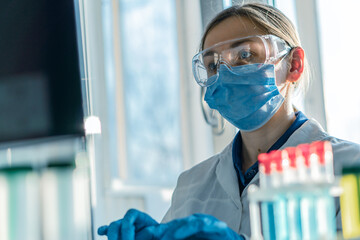 A female researcher sits at a workplace in a laboratory, behind a personal computer monitor. Against the background research statistics. Pharmaceutical medical worker in protection works at keyboard.