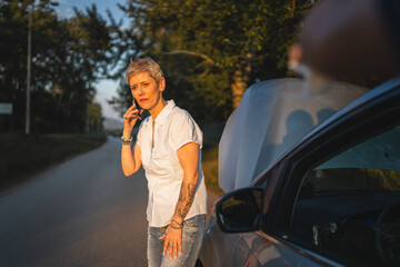 one woman mature female standing on the road in the evening sunset by the broken vehicle car automobile failed engine open hood making a phone call for help roadside assistance towing service concept