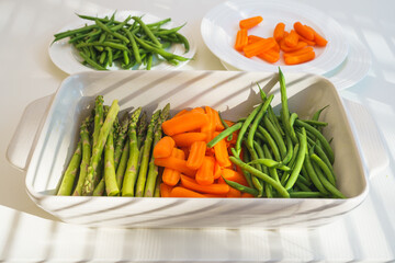 Raw organic green beans, baby carrots, green asparagus in baking pan, ready to be cooked. Close up view, white kitchen table background
